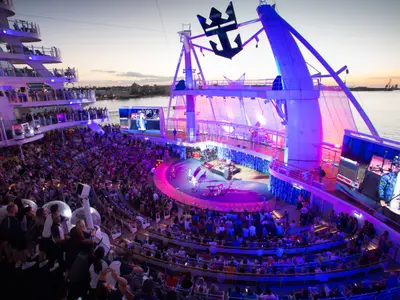 Abendliche Vorstellung im AquaTheater der Oasis of the Seas. Publikum auf den Rängen verfolgt Band und Tänzer auf der Bühne unter farbiger Beleuchtung. Große Bildschirme. Im Hintergrund Küste.