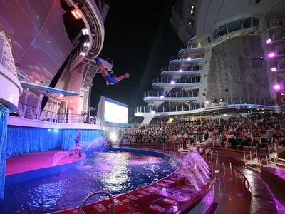 Nächtliche AquaTheater-Show auf der Oasis of the Seas. Ein Springer in der Luft, Künstler im Pool. Publikum auf Rängen. Schiffsfassade mit Kletterwand hinten.