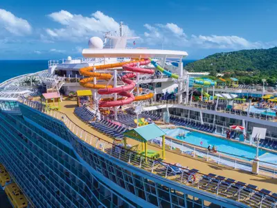 Luftaufnahme der Oasis of the Seas. Die bunten Wasserrutschen und Pools auf dem Oberdeck sind vor einer grünen Inselkulisse zu sehen.
