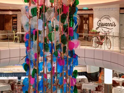 Atrium der Odyssey of the Seas mit großer bunter Kunstinstallation. Auf mehreren Ebenen sind Restaurants sichtbar, darunter Giovanni's mit rotem Vintage-Fahrrad.