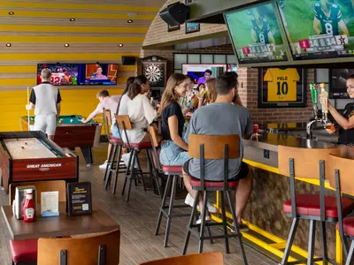 Eine moderne Sportsbar mit Holzvertäfelung und gelben Akzenten. Mehrere Personen spielen Billard und Shuffleboard, andere sitzen an der Bar, wo eine Barkeeperin ein Bier zapft. Bildschirme zeigen Sportübertragungen.