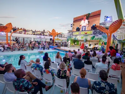 Auf der Odyssey of the Seas verfolgen Passagiere eine Pooldeck-Veranstaltung am Abend mit Bühne, Diskussionsrunde und großer Videowand. Obere Decks sind ebenfalls besetzt.