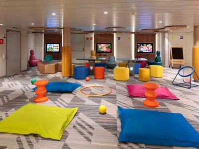 Helle und farbenfrohe Spielfläche an Bord der Odyssey of the Seas. Der Raum bietet Bildschirme, bunte Sitzsäcke, Hocker und diverse Spielgeräte auf gemustertem Teppich. Ein kinderfreundlicher Bereich.