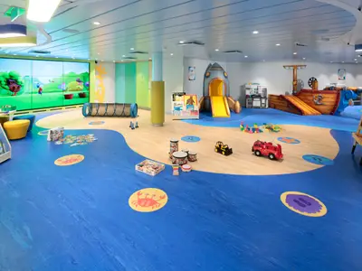 Heller Kinderspielbereich auf der Odyssey of the Seas mit Videowand, Raketenrutsche, Piratenschiff, Spieltunnel und diversen Spielzeugen auf blauem Boden.