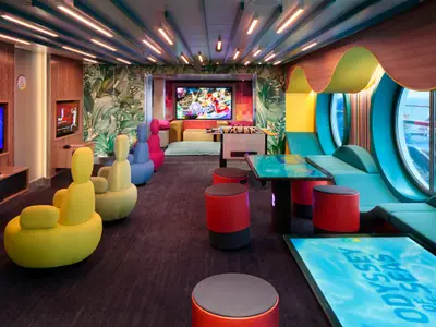 Bunter Freizeitbereich auf der Odyssey of the Seas mit Gaming-Stationen, weichen Sitzmöbeln, Kickertisch, großen Bullaugen und interaktivem Bodendisplay. Modernes Design.