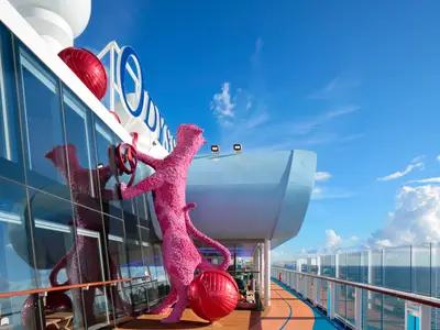 Auf der Odyssey of the Seas ist eine große pinke Tierskulptur mit rotem Rad an der Glasfassade zu sehen. Dahinter blaues Meer und Himmel mit Wolken.