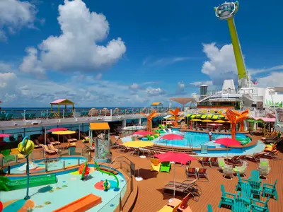 Das Sonnendeck der Odyssey of the Seas mit Hauptpool, farbenfrohem Kinderwasserpark, vielen Liegen und Sonnenschirmen. Im Hintergrund blaues Meer und Himmel. Ein gelb-grüner Beobachtungsarm ragt über das Schiff.