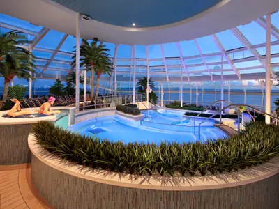 Abendliche Szene im Solarium der Odyssey of the Seas: Beleuchtete Pools, eine Frau am Beckenrand und Palmen unter einem Glasdach mit Meerblick.
