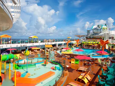 Sonniges Oberdeck der Odyssey of the Seas mit großem Pool, buntem Kinder-Wasserpark, Liegestühlen, Schirmen und der North Star Kapsel. Meer im Hintergrund.
