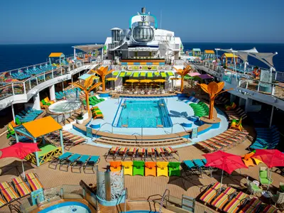Sonniges Oberdeck der Odyssey of the Seas mit Pools, bunten Liegestühlen, Whirlpools und der markanten North Star Kapsel. Dahinter ist das blaue Meer zu sehen.
