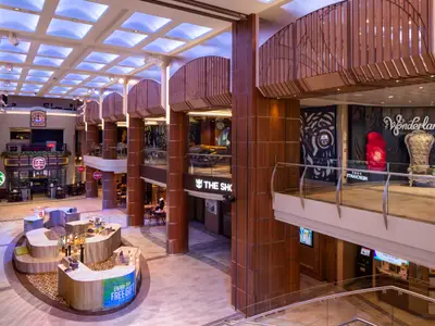 Weitwinkelaufnahme des mehrstöckigen Atriums an Bord der Odyssey of the Seas. Zu sehen sind Starbucks, ein Pub, Geschäfte und das Wonderland Restaurant. Helle Deckenbeleuchtung und Holzverkleidungen prägen den Raum.