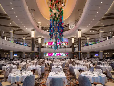 Innenansicht des Hauptrestaurants der Odyssey of the Seas. Viele gedeckte Tische, dazu Sitzbereiche auf oberen Ebenen. Ein großer, bunter Kronleuchter und Bildschirme mit Kunst schmücken den Raum.