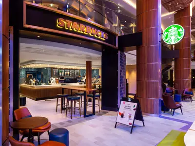 Das Starbucks-Café an Bord der Odyssey of the Seas mit Eingang, Theke, Menütafeln und Sitzgelegenheiten im modernen Interieur des Schiffs.