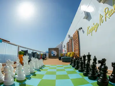 Ein sonniges Außendeck der Odyssey of the Seas zeigt ein großes Schachbrett mit weißen und schwarzen Figuren. Sitzbereiche und das offene Meer sind im Hintergrund sichtbar.