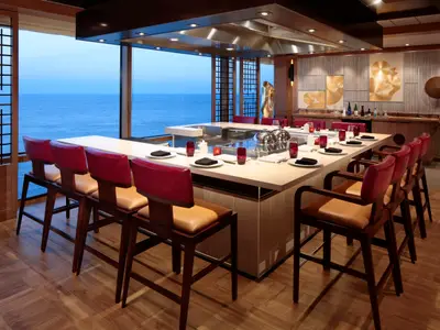 Teppanyaki-Restaurant auf der Odyssey of the Seas. Lange Theke mit roten Barhockern. Meerblick durch Fenster. Moderne goldene Kunst.