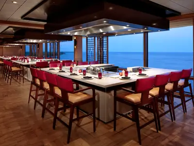 Teppanyaki-Restaurant der Odyssey of the Seas: Lange Tische mit roten Barhockern, Blick auf den Ozean bei Dämmerung.