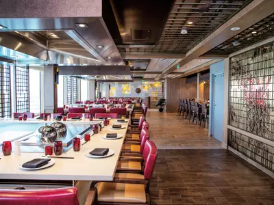 Das Teppanyaki-Restaurant der Odyssey of the Seas. Lange Tische mit Kochplatten, rote Stühle und eine Bar. Modernes Design mit großen Fenstern und asiatischen Akzenten.