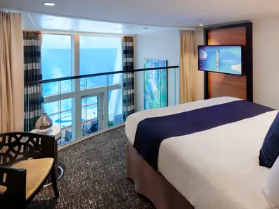 Helle Loft-Kabine auf der Odyssey of the Seas. Man sieht ein Bett, TV, Stuhl und Panoramafenster mit Meerblick und einem unteren Balkon.