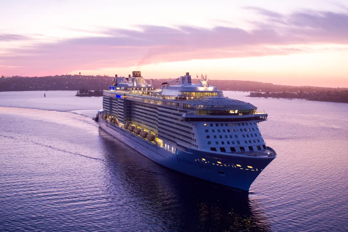 Die moderne Ovation of the Seas fährt bei Sonnenuntergang auf ruhigem Wasser, mit einer Stadt-Skyline im Hintergrund. Der Himmel ist in sanften Lila- und Orangetönen gefärbt.
