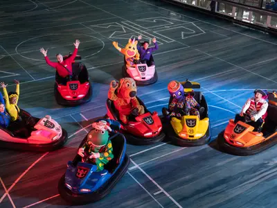 Auf der Ovation of the Seas sind mehrere Personen, darunter die Wiggles und ihre Maskottchen, in bunten Bumper-Cars auf einer Indoor-Fläche zu sehen. Sie fahren und winken fröhlich.