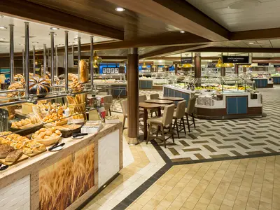 Das Buffetrestaurant der Ovation of the Seas zeigt eine reichhaltige Brottheke im Vordergrund, dahinter mehrere Essensstationen mit frischen Speisen und Sitzgelegenheiten für Gäste.