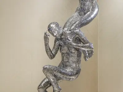 Silberne Skulptur einer geflügelten Figur in nachdenklicher Pose. Gefertigt aus komplexen, netzartigen Mustern vor heller Wand.