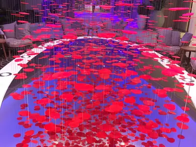Eine faszinierende Kunstinstallation mit Hunderten schwebender roter Scheiben ist auf der Ovation of the Seas zu sehen. Die Scheiben hängen über einem stark reflektierenden, blau beleuchteten Boden, der ihre Schatten und die umgebende, leicht unscharfe Lounge-Sitzgruppe widerspiegelt.