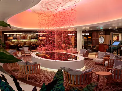 Innenansicht der Ovation of the Seas: Eine elegante Lounge mit roter Deckenbeleuchtung und einem ovalen Wasserbecken, umgeben von Holzstühlen. Im Hintergrund sind die VINTAGES Bar und das Restaurant Jamie's Italian zu sehen.