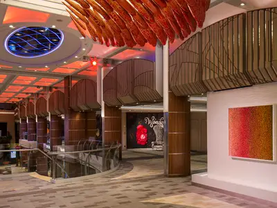 Blick ins Atrium der Ovation of the Seas: Rote Deckeninstallation, blaue Beleuchtung, Holzsäulen, Glasbrüstung. 'Wonderland'-Wand und Kunstwerk sichtbar.