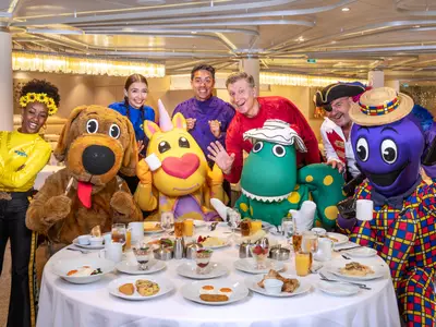 Ein fröhliches Frühstück mit den Wiggles und ihren Maskottchen an Bord der Ovation of the Seas. Mehrere Personen und Kostümfiguren sitzen um einen gedeckten Tisch mit Speisen.