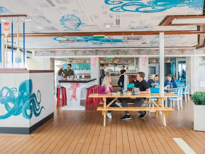 Der belebte Restaurantbereich auf der Ovation of the Seas zeigt Gäste an Tischen und einer Bar. Maritimes Dekor mit blauen Tintenfischen, Paddeln und Fischmotiven prägt den Raum.