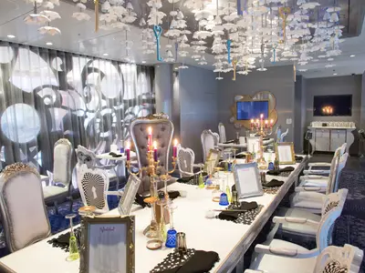 Langes, weiß gedecktes Restaurant mit verschiedenen Stühlen und Kerzenleuchtern. An der Decke hängen weiße Blüten und goldene Schlüssel. Im Hintergrund Fenster mit Mustern, zwei Bildschirme und ein Sideboard.