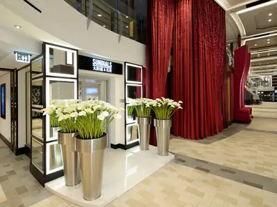 Blick auf einen eleganten Innenbereich der Ovation of the Seas mit silbernen Blumenkübeln, einem Uhrengeschäft, roten Samtvorhängen und einem breiten Gang.