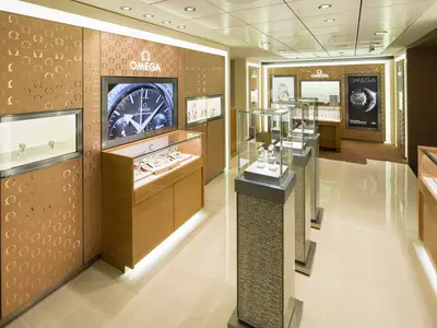 Innenansicht der luxuriösen Omega Boutique an Bord der Ovation of the Seas. Zu sehen sind helle Wände mit Omega-Logo, Glasvitrinen mit Uhren, ein großer Bildschirm und eine helle Bodenfläche.