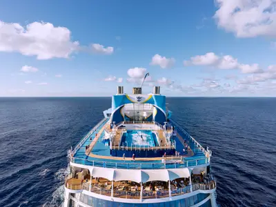 Das Heck der Ovation of the Seas auf hoher See. Der FlowRider-Surfsimulator und Passagiere auf den Decks sind sichtbar. Blauer Himmel und weites Meer.