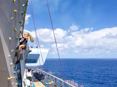 Eine junge Person klettert die Wand der Ovation of the Seas empor. Zu sehen ist die Kletterin mit Gurt und das blaue Meer mit Wolken im Hintergrund.