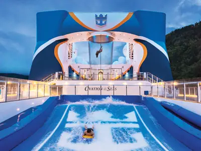 Zwei Personen nutzen Sportattraktionen auf dem Außendeck der Ovation of the Seas. Eine fliegt im Skydiving-Simulator, eine liegt auf dem Surfsimulator. Hinten ein bewaldeter Berg.