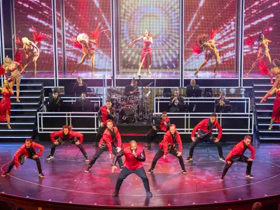Bühnenshow an Bord der Ovation of the Seas: Männliche Tänzer und Sänger in Rot, eine Live-Band und weibliche Tänzerinnen mit Federflügeln auf gestuften Bühnen vor LED-Bildschirmen.