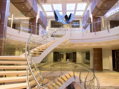 Blick auf das mehrstöckige Atrium der Quantum of the Seas mit einer geschwungenen Holztreppe, Glasgeländern und einer blauen, wellenförmigen Kunstinstallation an der Decke.
