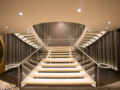 Moderne, beleuchtete Doppeltreppe mit Glas- und Edelstahlgeländer im Interieur der Quantum of the Seas. Die Umgebung zeigt vertikale Wandverkleidungen und eine helle Decke.