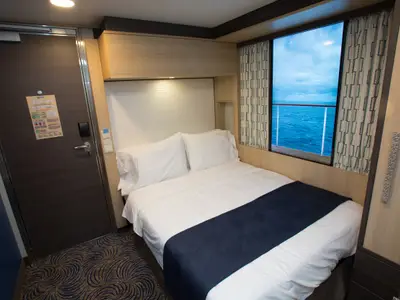 Innenansicht einer hellen Kabine an Bord der Quantum of the Seas. Zu sehen sind ein gemachtes Bett, ein großes Fenster mit Meerblick, ein Kleiderschrank und die Kabinentür mit Informationsblatt.