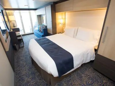 Moderne Kabine auf der Quantum of the Seas. Ein großes Bett mit weißer Bettwäsche und blauem Läufer, Schreibtisch, blaues Sofa und Balkontür sind sichtbar. Der Boden hat einen gemusterten blauen Teppich.