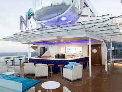 Eine moderne Außenbar auf dem Oberdeck der Quantum of the Seas mit blau beleuchtetem Tresen, Loungemöbeln und Meerblick. Darüber schwebt die futuristische North Star Kapsel.