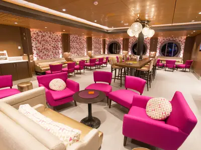 Moderne Lounge auf der Quantum of the Seas. Helle Einrichtung mit pinken Sesseln, beigen Sofas, geometrischer Tapete, runden Fenstern und einem langen Holztisch mit Barhockern.