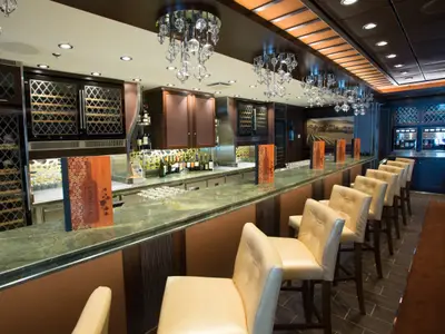 Innenansicht der modernen Vintages Weinbar auf der Quantum of the Seas. Eine lange Theke mit grüner Marmorplatte und beigen Barhockern ist zu sehen, im Hintergrund Weinflaschen und ein Weinberg-Bild.
