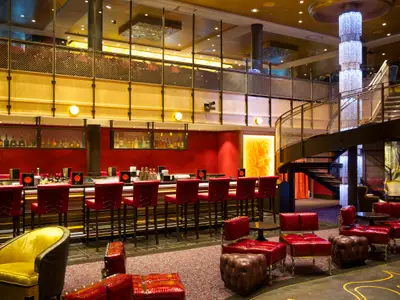 Elegante Bar auf der Quantum of the Seas: Rote Theke mit Hockern, Wendeltreppe zur Empore und großer Kristallleuchter. Rote und braune Sitzgruppen füllen den Raum.