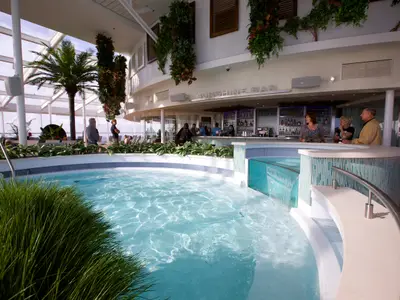 Ein heller Solarium-Bereich an Bord der Quantum of the Seas mit einem runden Pool und der dahinterliegenden Sunshine Bar. Mehrere Gäste entspannen oder stehen an der Bar, während Tageslicht durch das Glasdach fällt und üppige Pflanzen den Raum schmücken.