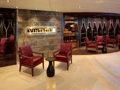 Loungebereich der Quantum of the Seas mit roten Sesseln und dunklen Tischen. Die Vintages Weinbar mit Steinwand und Flaschen-Dekor ist im Hintergrund zu sehen. Einladende Atmosphäre.