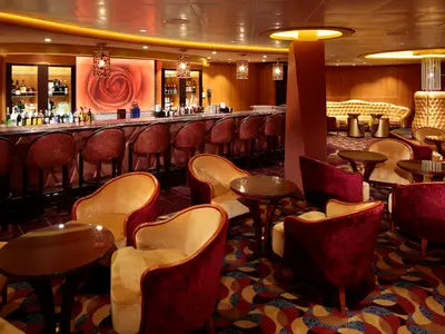 Bar- und Loungebereich der Quantum of the Seas. Theke mit Flaschen, Barhockern, ein großes Rosenbild. Sessel, Tische, gemusterter Teppich in warmen Farben.