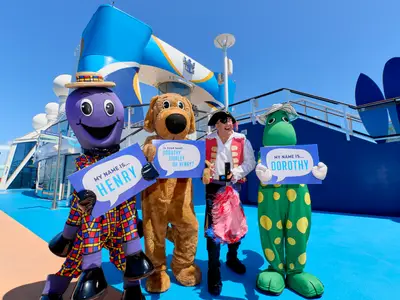 Wiggles-Charaktere Henry, Wags, Captain Feathersword und Dorothy posieren auf dem sonnigen Deck der Quantum of the Seas und halten Schilder.